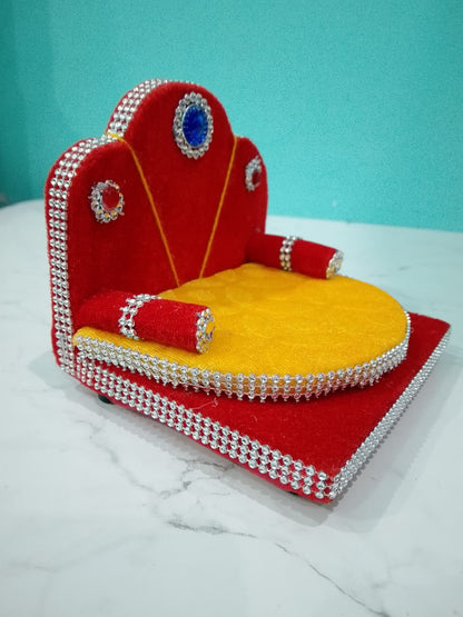 Singhasan for Gopal ji with red and yellow cushions on a white surface with teal background