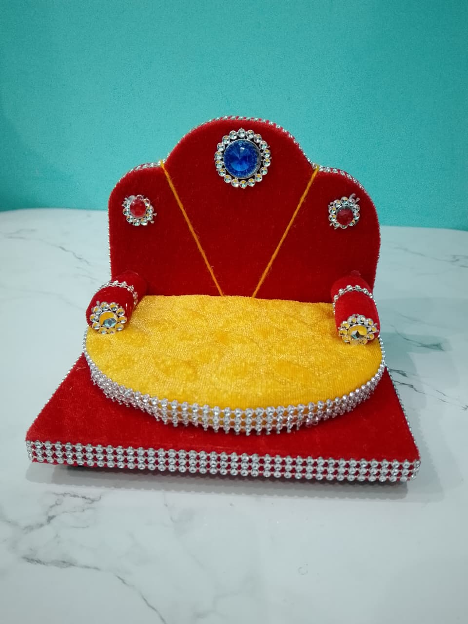 Singhasan for Gopal ji with red and yellow cushions on a white surface with teal background