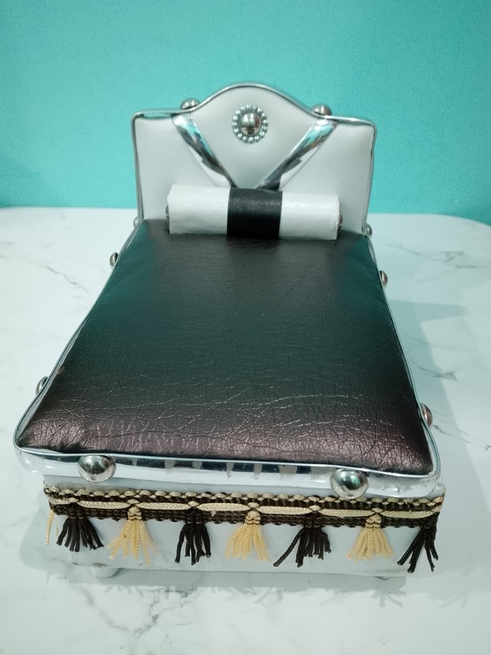 Silver Bed for Laddu Gopal Radha Krishna with black leather top and tassels on a white surface with teal background