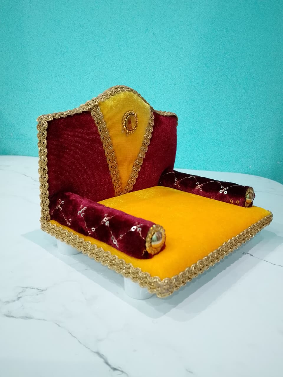 Singhasan for Gopal ji with red and yellow cushions on a white surface with teal background