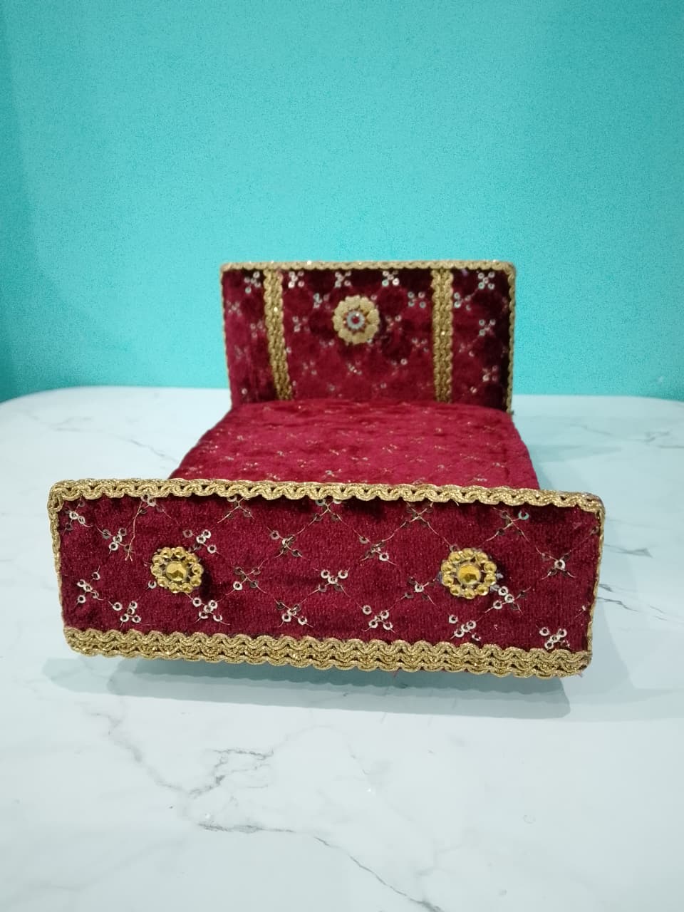 Red velvet bed with gold trim on a white surface against a teal background for Laddu gopal and Radha Krishna