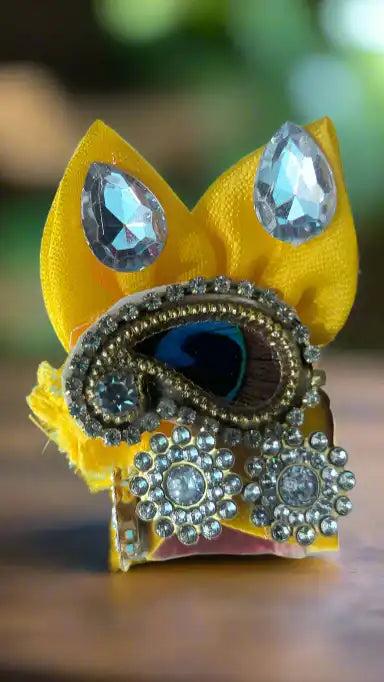 Laddu Gopal pagdi with yellow fabric and metallic embellishments on a blurred background